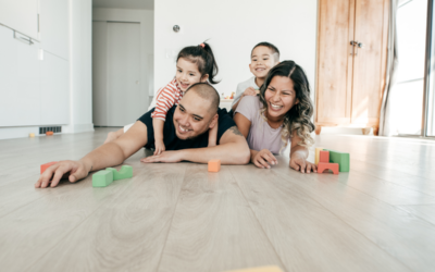 A happy family, including a mother, a father, and two young children, plays together on a light colored hardwood floor in their home.