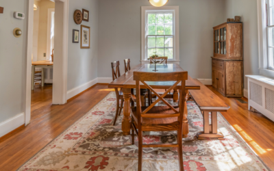 A dining room with a traditional patterned area rug beneath a wooden dining table, seated for six with four chairs and a bench, atop hardwood flooring.