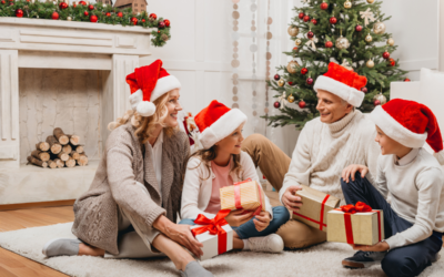 A smiling family of four in Santa hats exchanges Christmas gifts while seated on a cozy rug over hardwood floors, with a decorated Christmas tree in the background.