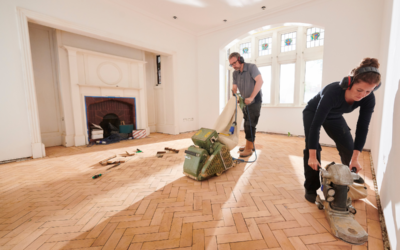 A man and woman using belt sanders to refinish herringbone wood floors in a bright room, but they forgot to wear dust masks.