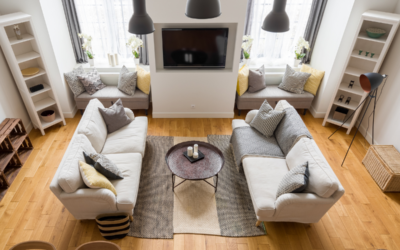 An aerial view of a living room with two beige couches facing each other, a modern fireplace with a TV mounted above it, and blonde hardwood floors illuminated by natural light.