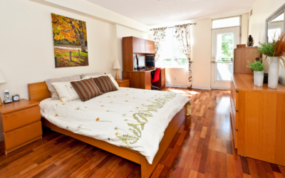 A cozy bedroom featuring midcentury modern wood furniture that matches the polished hardwood floors, illuminated by natural light from large windows.